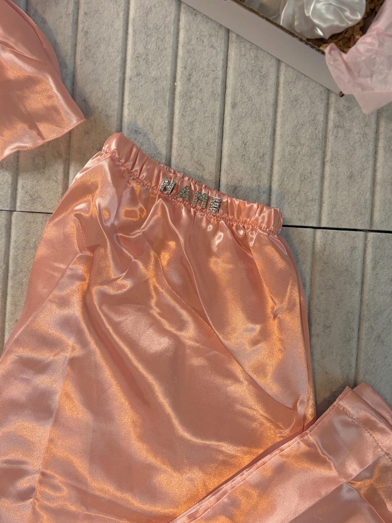 Op de afbeelding: Perzikroze satijnen pyjamaset met het woord "NAME" in zilveren strass-steentjes op de tailleband. De set bevat een broek en een bijpassende slaapmuts. De stof heeft een glanzende, gladde textuur.