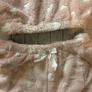 K&ouml;nnte beinhalten: Ein zweiteiliger Pyjama-Set aus weichem, pl&uuml;schigem Material. Das Set ist hellrosa mit wei&szlig;en herzf&ouml;rmigen Mustern. Das Oberteil ist im Cropped-Stil gehalten, und die Hose hat eine wei&szlig;e Schleife mit Strass-Akzent an der Taille. Der Stoff wirkt flauschig und gem&uuml;tlich.