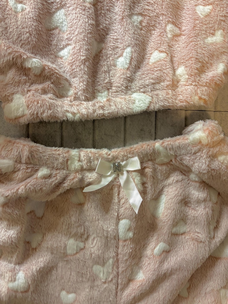 K&ouml;nnte beinhalten: Ein zweiteiliger Pyjama-Set aus weichem, pl&uuml;schigem Material. Das Set ist hellrosa mit wei&szlig;en herzf&ouml;rmigen Mustern. Das Oberteil ist im Cropped-Stil gehalten, und die Hose hat eine wei&szlig;e Schleife mit Strass-Akzent an der Taille. Der Stoff wirkt flauschig und gem&uuml;tlich.