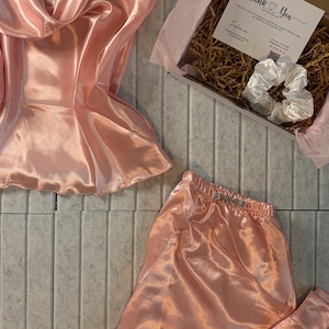 Op de afbeelding: Een perzikroze satijnen pyjamaset, bestaande uit een camisole top en shorts, wordt getoond. Een geschenkdoos met een witte scrunchie en een kaart met "Thank You" is ook zichtbaar. De set is geplaatst op een lichtgrijze betegelde ondergrond.