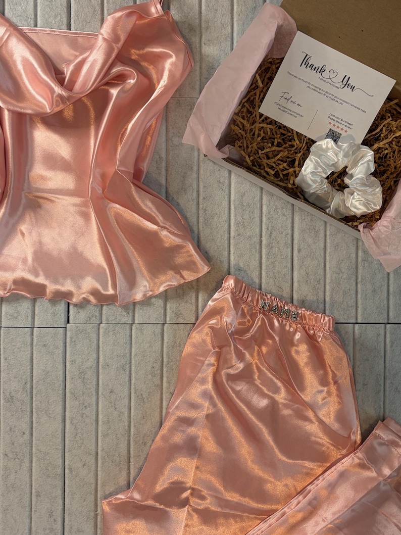 Op de afbeelding: Een perzikroze satijnen pyjamaset, bestaande uit een camisole top en shorts, wordt getoond. Een geschenkdoos met een witte scrunchie en een kaart met "Thank You" is ook zichtbaar. De set is geplaatst op een lichtgrijze betegelde ondergrond.