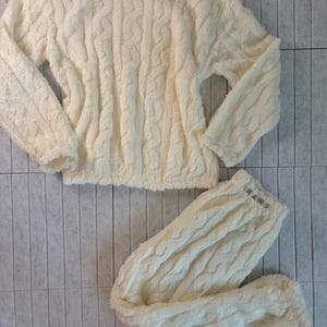 May include: Cream-colored, cable-knit pajama set. The set includes a long-sleeved top and matching pants. The fabric appears soft and plush, with a textured, woven pattern. The pants have the word "MARS" printed on the waistband.