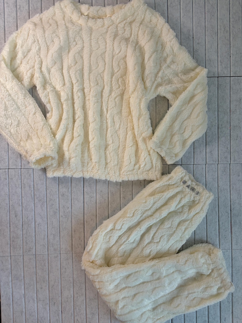 May include: Cream-colored, cable-knit pajama set. The set includes a long-sleeved top and matching pants. The fabric appears soft and plush, with a textured, woven pattern. The pants have the word "MARS" printed on the waistband.