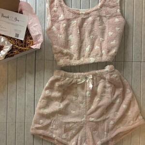 K&ouml;nnte beinhalten: Ein rosa, flauschiges Pyjama-Set mit wei&szlig;en Herzmustern. Das Set besteht aus einem Crop-Top und Shorts, beide mit einem weichen, strukturierten Aussehen. Eine wei&szlig;e Schleife ziert die Shorts. Eine Geschenkbox mit einer "Thank You"-Karte ist sichtbar.