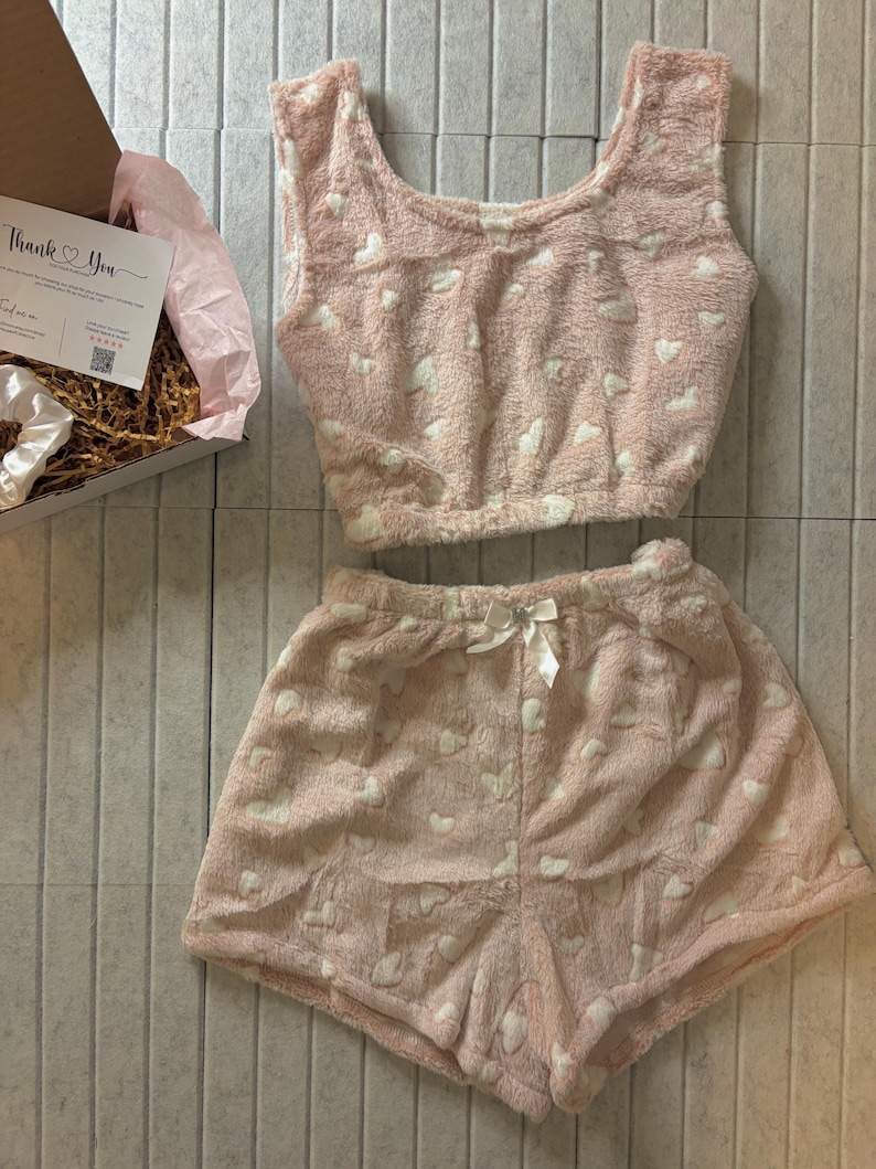 K&ouml;nnte beinhalten: Ein rosa, flauschiges Pyjama-Set mit wei&szlig;en Herzmustern. Das Set besteht aus einem Crop-Top und Shorts, beide mit einem weichen, strukturierten Aussehen. Eine wei&szlig;e Schleife ziert die Shorts. Eine Geschenkbox mit einer "Thank You"-Karte ist sichtbar.