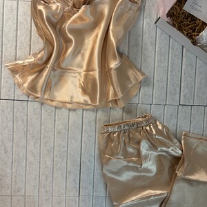 Op de afbeelding: Een champagnekleurige satijnen pyjamaset. De set bevat een camisole top met een gefranjerde zoom en een bijpassende pyjamabroek. De stof heeft een glanzende glans en de elastische tailleband is zichtbaar op de broek. De set wordt gepresenteerd op een lichtgrijze ondergrond.