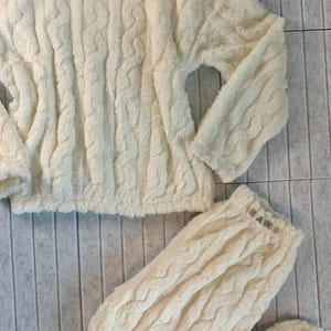 May include: Cream-colored, cable-knit pajama set. The set includes a long-sleeved top and matching pants. The pants have the word "HANG" embroidered on the waistband. The fabric has a soft, plush texture.