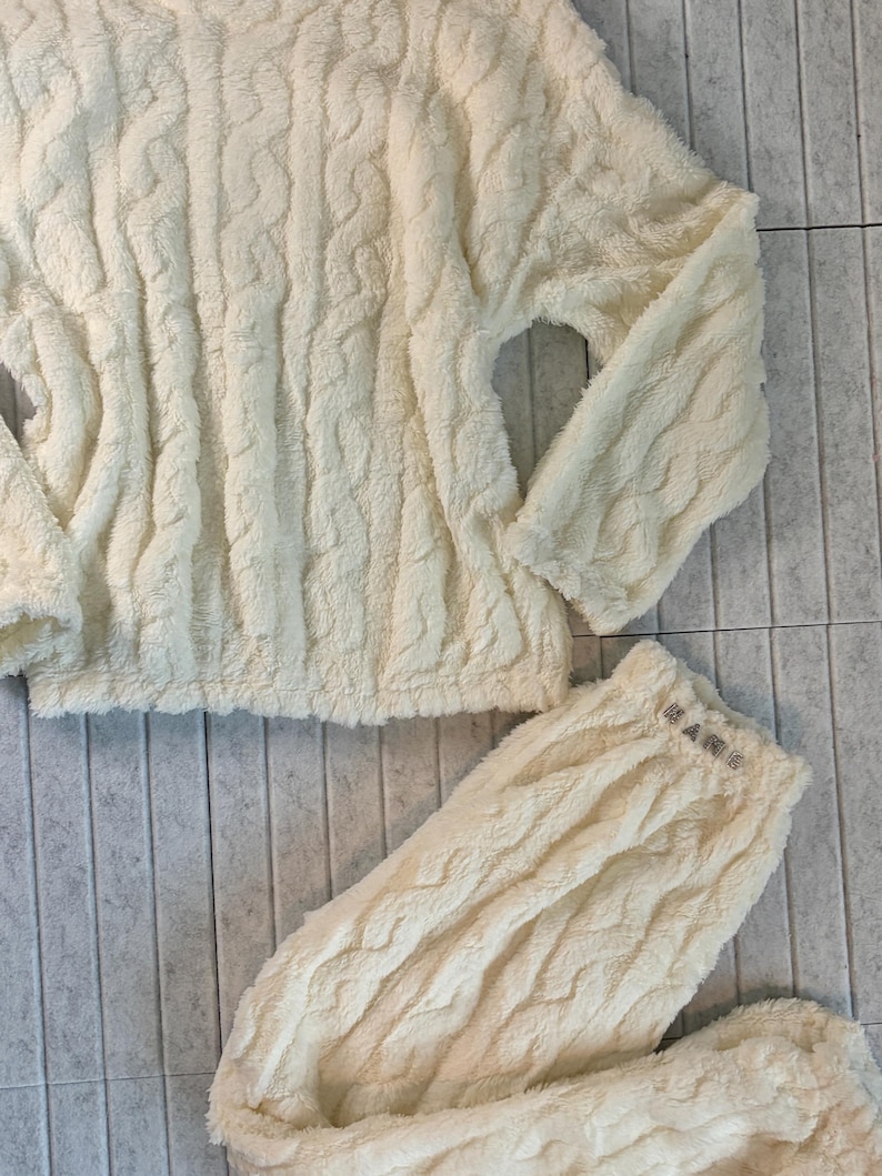 May include: Cream-colored, cable-knit pajama set. The set includes a long-sleeved top and matching pants. The pants have the word "HANG" embroidered on the waistband. The fabric has a soft, plush texture.