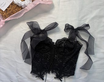 May include: Black lace corset top with sheer ribbon shoulder straps and small bows. The top is on a white surface, with a pink box containing a white scrunchie and a card in the background.