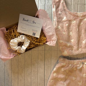 K&ouml;nnte beinhalten: Ein rosa, flauschiges Pyjama-Set mit wei&szlig;en Herzmustern und einem passenden Oberteil. Ein wei&szlig;es Satin-Haargummi und eine Karte mit der Aufschrift "Thank You" befinden sich in einer Pappschachtel mit dekorativem Stroh.