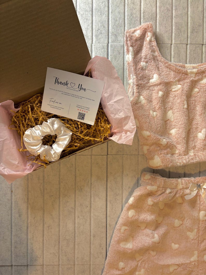 K&ouml;nnte beinhalten: Ein rosa, flauschiges Pyjama-Set mit wei&szlig;en Herzmustern und einem passenden Oberteil. Ein wei&szlig;es Satin-Haargummi und eine Karte mit der Aufschrift "Thank You" befinden sich in einer Pappschachtel mit dekorativem Stroh.