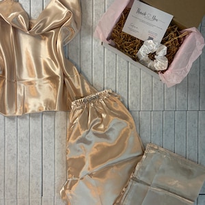 May include: A champagne-colored satin pajama set, including a camisole top and pants, is displayed. A gift box with a "Thank You" card and a white scrunchie is also present. The set is arranged on a light gray tiled surface.