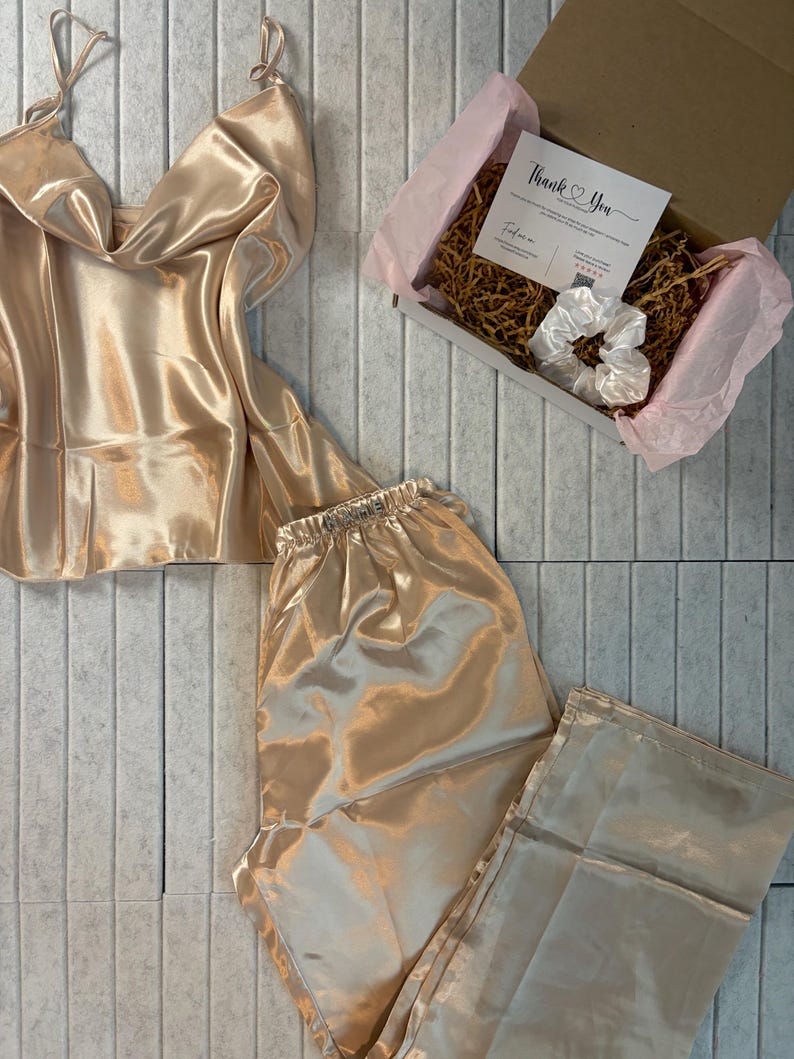 May include: A champagne-colored satin pajama set, including a camisole top and pants, is displayed. A gift box with a "Thank You" card and a white scrunchie is also present. The set is arranged on a light gray tiled surface.