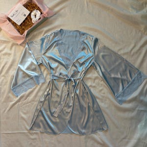May include: A light blue satin robe featuring wide sleeves with lace trim at the cuffs and along the V-neckline. The robe includes a matching fabric tie belt at the waist. A gift box containing a white scrunchie and a thank you card is nearby.