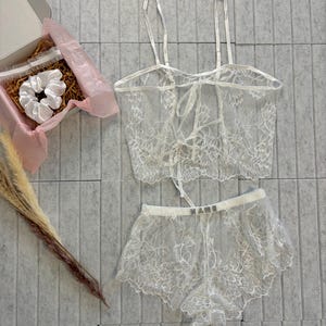 Puede incluir: Un conjunto de lencería de encaje blanco con top y shorts a juego. El top tiene tirantes finos y espalda atada. Los shorts tienen la palabra "NAME" en la cintura. También se incluye una caja de regalo con una goma para el pelo.