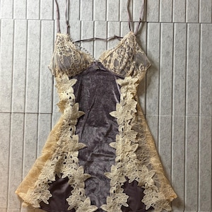 May include: A sleeveless chemise featuring a dark gray velvet center panel and beige lace cups. The garment is accented with intricate floral lace trim along the sides and thin adjustable shoulder straps, creating a layered appearance.