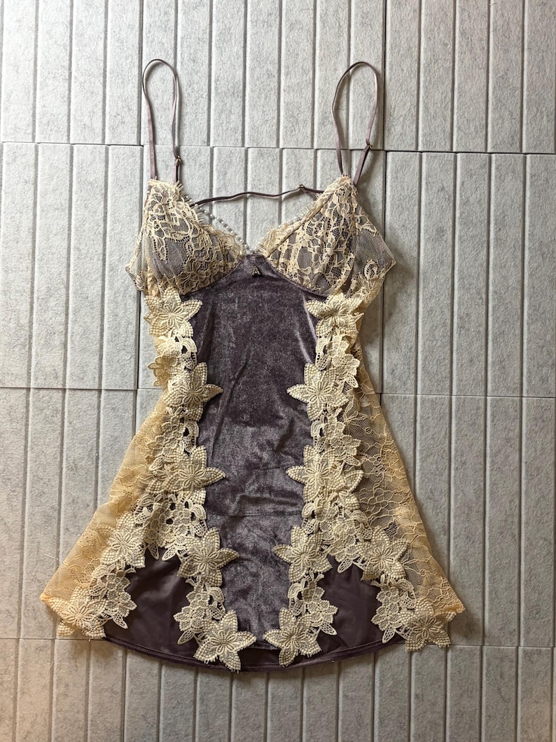 May include: A sleeveless chemise featuring a dark gray velvet center panel and beige lace cups. The garment is accented with intricate floral lace trim along the sides and thin adjustable shoulder straps, creating a layered appearance.
