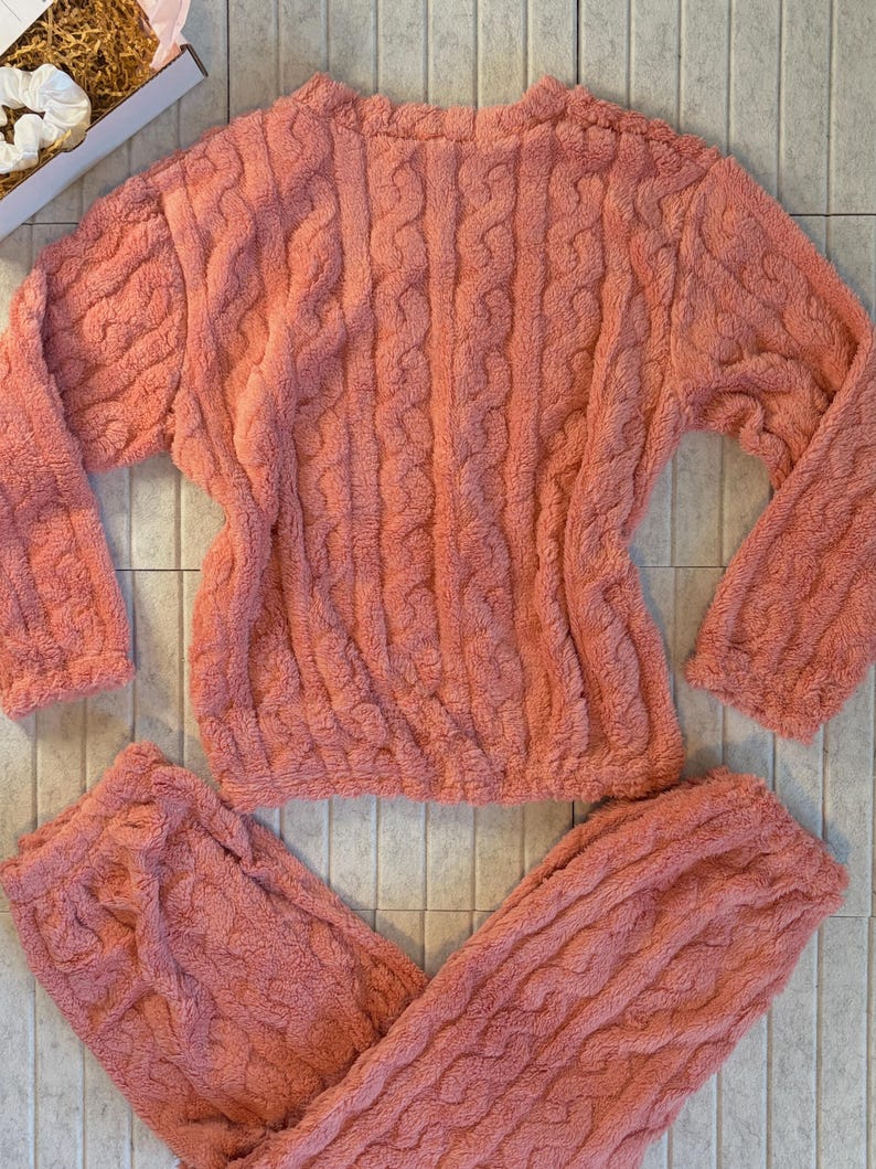 May include: A coral-colored, plush pajama set featuring a cable knit design. The set includes a long-sleeved top and matching pants. The fabric appears soft and cozy, perfect for lounging. The set is displayed on a tiled surface.