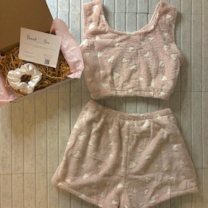 K&ouml;nnte beinhalten: Ein rosa, flauschiges Pyjama-Set mit wei&szlig;en Herzmustern, bestehend aus einem Crop-Top und Shorts. Eine Box mit einer Dankeskarte und einem wei&szlig;en Scrunchie ist ebenfalls sichtbar.