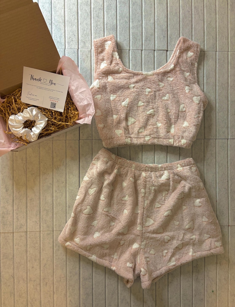 K&ouml;nnte beinhalten: Ein rosa, flauschiges Pyjama-Set mit wei&szlig;en Herzmustern, bestehend aus einem Crop-Top und Shorts. Eine Box mit einer Dankeskarte und einem wei&szlig;en Scrunchie ist ebenfalls sichtbar.