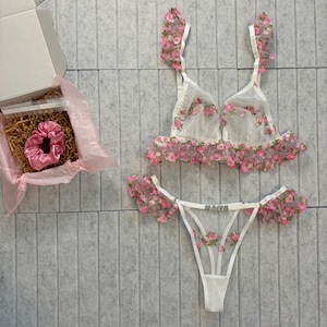 May include: A white lingerie set with pink floral embroidery, including a bra and thong. A pink scrunchie is in an open box with pink tissue paper. The thong has the word "NAME" on it.