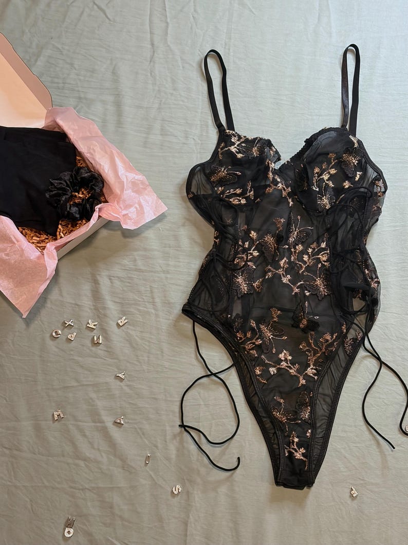 May include: Black lingerie set with floral embroidery. The set includes a sheer bodysuit with adjustable straps and a high-cut leg, along with a matching pair of underwear and a scrunchie, presented in a gift box.