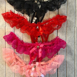 May include: Four pairs of lingerie in black, red, fuchsia, and light pink. Each pair features lace trim, satin bows, and decorative embellishments. The lingerie is displayed against a neutral background.