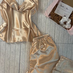 May include: A champagne-colored satin pajama set, including a camisole top and shorts, is displayed. A gift box with a "Thank You" card and a white scrunchie is also visible. The set is made of a shiny, smooth fabric.