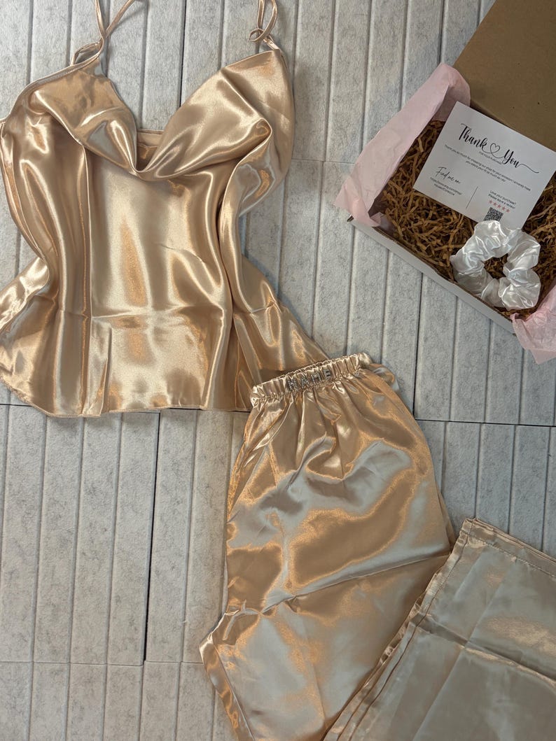 May include: A champagne-colored satin pajama set, including a camisole top and shorts, is displayed. A gift box with a "Thank You" card and a white scrunchie is also visible. The set is made of a shiny, smooth fabric.