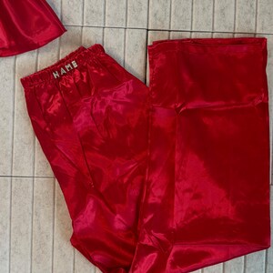 May include: Bright red satin pajama pants with an elastic waistband. The waistband features the word "NAME" in silver letters. The pants are part of a set, with a matching top visible in the image.
