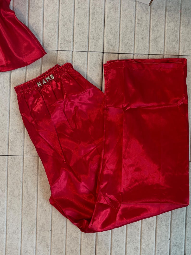 May include: Bright red satin pajama pants with an elastic waistband. The waistband features the word "NAME" in silver letters. The pants are part of a set, with a matching top visible in the image.