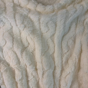 May include: Close-up of a cream-colored, plush fabric with a textured, cable-knit design. The soft material has a thick, furry appearance, with vertical ridges and a slightly ruffled edge. The overall look is cozy and inviting.