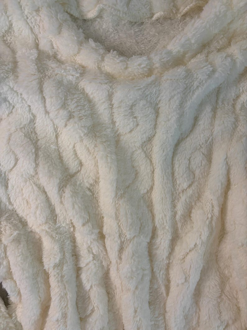 May include: Close-up of a cream-colored, plush fabric with a textured, cable-knit design. The soft material has a thick, furry appearance, with vertical ridges and a slightly ruffled edge. The overall look is cozy and inviting.