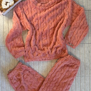 May include: A coral-colored, cable-knit pajama set. The set includes a long-sleeved top and matching pants. The top has a crew neck and the pants have a drawstring waist. The fabric appears soft and plush, perfect for lounging.