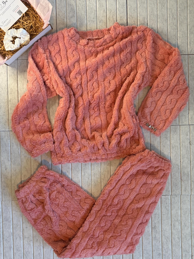 May include: A coral-colored, cable-knit pajama set. The set includes a long-sleeved top and matching pants. The top has a crew neck and the pants have a drawstring waist. The fabric appears soft and plush, perfect for lounging.