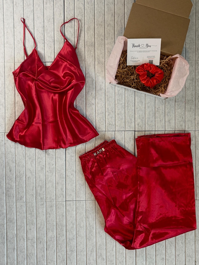May include: A red satin pajama set with a camisole top and matching pants. The set includes a gift box with a scrunchie and a card that says "Thank You". The pajama pants have the word "NAME" embroidered on the waistband.