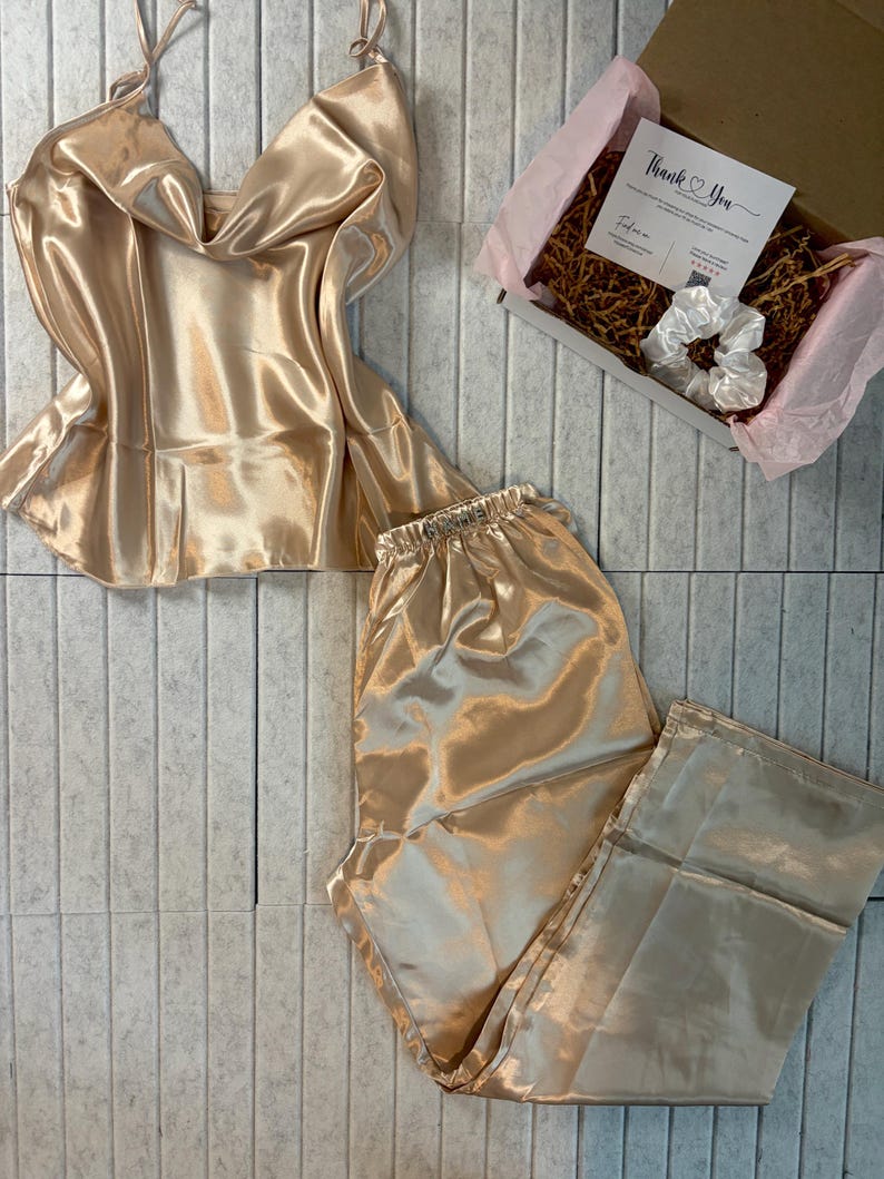 May include: A champagne-colored satin pajama set, including a camisole top and pants, is displayed. A gift box with a "Thank You" card and a white scrunchie is also visible. The set is arranged on a light gray, textured surface.