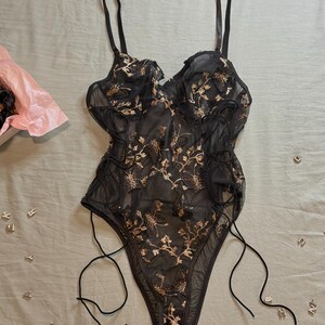 May include: Black lingerie bodysuit with gold floral embroidery. The sheer fabric features a delicate pattern, adjustable straps, and side ties. A pink tissue-lined box is visible in the background, along with scattered silver letter charms.
