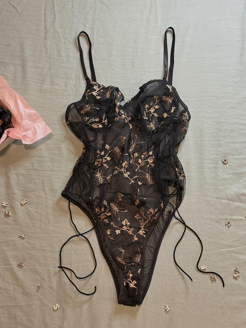 May include: Black lingerie bodysuit with gold floral embroidery. The sheer fabric features a delicate pattern, adjustable straps, and side ties. A pink tissue-lined box is visible in the background, along with scattered silver letter charms.