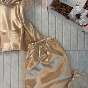 Op de afbeelding: Een luxe champagnekleurige satijnen pyjamaset, bestaande uit een camisole top en bijpassende broek. De stof heeft een zachte glans. Een witte scrunchie en een doos met een bedankkaart zijn ook zichtbaar.