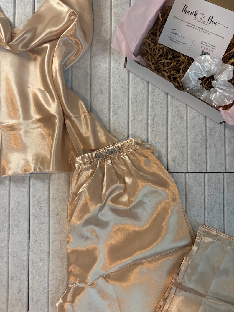 Op de afbeelding: Een luxe champagnekleurige satijnen pyjamaset, bestaande uit een camisole top en bijpassende broek. De stof heeft een zachte glans. Een witte scrunchie en een doos met een bedankkaart zijn ook zichtbaar.