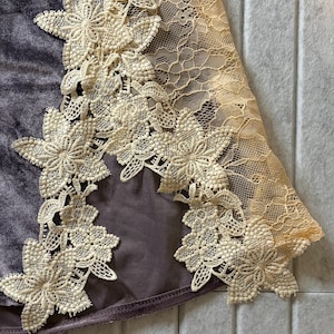 May include: A dark purple velvet fabric panel featuring an intricate cream-colored floral lace applique along the edge. The lace pattern consists of detailed flower motifs connected by a delicate mesh netting, contrasting against the smooth texture.