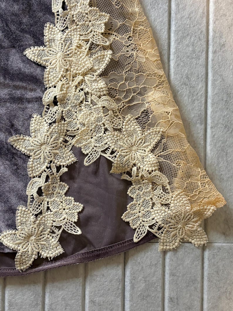 May include: A dark purple velvet fabric panel featuring an intricate cream-colored floral lace applique along the edge. The lace pattern consists of detailed flower motifs connected by a delicate mesh netting, contrasting against the smooth texture.