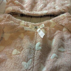 K&ouml;nnte beinhalten: Ein zweiteiliges rosa Pl&uuml;sch-Pyjama-Set mit wei&szlig;en Herzmustern. Ober- und Unterteil bestehen aus einem weichen, flauschigen Material. Eine wei&szlig;e Schleife mit dekorativer Mitte befindet sich auf der Vorderseite der Pyjamahose.