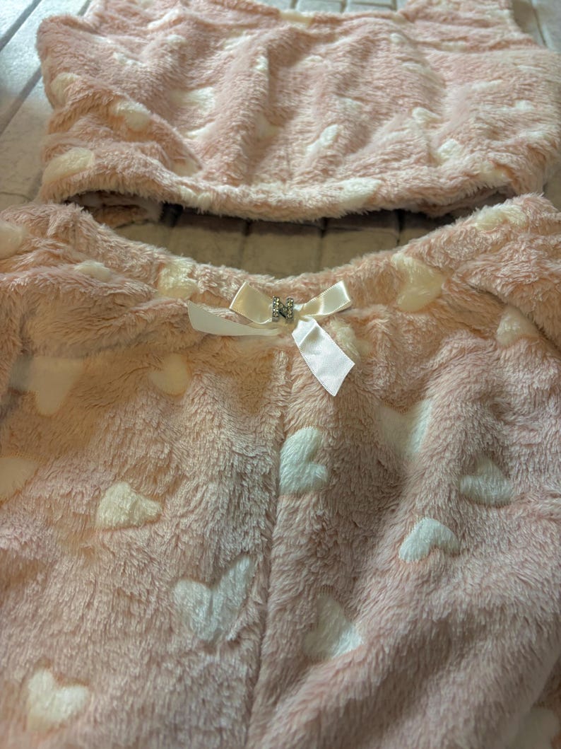 K&ouml;nnte beinhalten: Ein zweiteiliges rosa Pl&uuml;sch-Pyjama-Set mit wei&szlig;en Herzmustern. Ober- und Unterteil bestehen aus einem weichen, flauschigen Material. Eine wei&szlig;e Schleife mit dekorativer Mitte befindet sich auf der Vorderseite der Pyjamahose.