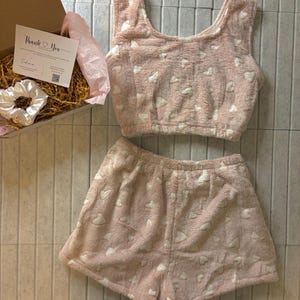 K&ouml;nnte beinhalten: Ein rosa, flauschiges Pyjama-Set mit wei&szlig;en Herzmustern. Das Set beinhaltet ein Crop-Top und Shorts. Eine Geschenkbox mit einer "Thank You"-Karte und einem wei&szlig;en Scrunchie ist ebenfalls sichtbar.