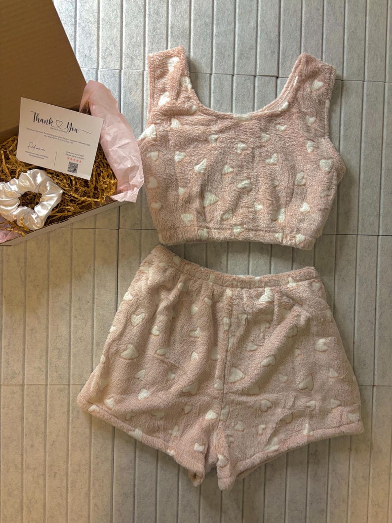 K&ouml;nnte beinhalten: Ein rosa, flauschiges Pyjama-Set mit wei&szlig;en Herzmustern. Das Set beinhaltet ein Crop-Top und Shorts. Eine Geschenkbox mit einer "Thank You"-Karte und einem wei&szlig;en Scrunchie ist ebenfalls sichtbar.