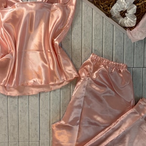 Op de afbeelding: Een lichtroze satijnen pyjamaset, bestaande uit een camisole top en een broek, wordt getoond op een lichtgrijze ondergrond. Een witte scrunchie en een kleine geschenkdoos met een kaart zijn ook zichtbaar. De set heeft een glanzende, gladde textuur.