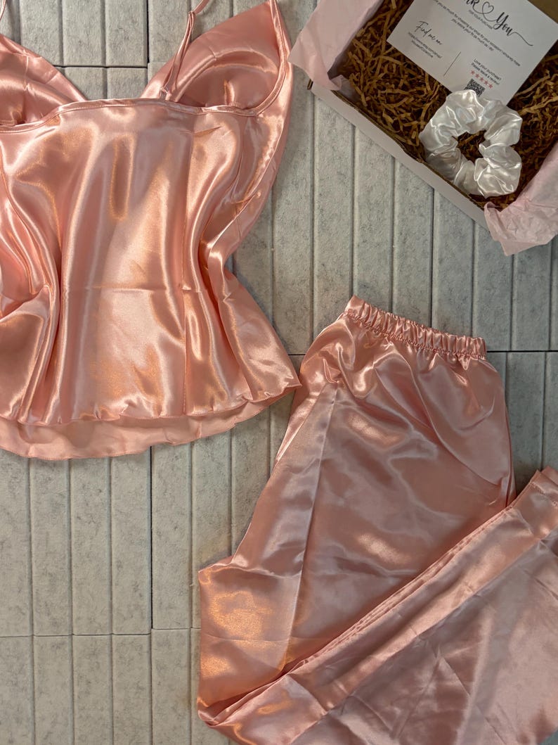 Op de afbeelding: Een lichtroze satijnen pyjamaset, bestaande uit een camisole top en een broek, wordt getoond op een lichtgrijze ondergrond. Een witte scrunchie en een kleine geschenkdoos met een kaart zijn ook zichtbaar. De set heeft een glanzende, gladde textuur.