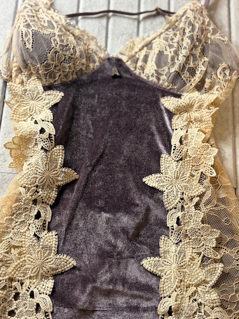 May include: A dark purple velvet bodysuit featuring cream-colored lace panels along the sides and a matching lace bodice. The garment includes thin adjustable straps and intricate floral lace appliques that frame the central velvet torso section.